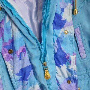 May include: A light blue jacket with a zipper closure and a purple and blue abstract print. The jacket has a hood and a pocket on the right side.