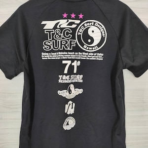 May include: Black t-shirt with a white graphic print of a yin-yang symbol and the text "T&C Surf Designs Hawaii". The shirt also features the text "Being a local at Makaha beach on the West side of Oahu He tuned his skills by offering custom shapes to his friends. Word got out fast... hence the move to open a store front that would house the custom shapes 71# T&C Surf Designs Hawaii".