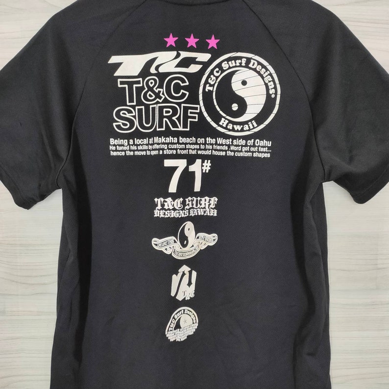 May include: Black t-shirt with a white graphic print of a yin-yang symbol and the text "T&C Surf Designs Hawaii". The shirt also features the text "Being a local at Makaha beach on the West side of Oahu He tuned his skills by offering custom shapes to his friends. Word got out fast... hence the move to open a store front that would house the custom shapes 71# T&C Surf Designs Hawaii".
