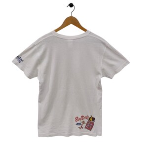 May include: White t-shirt with short sleeves, hanging on a wooden hanger. The shirt features a graphic on the lower right that says "Surf Days" with a cartoon character and a chocolate bar. The left sleeve has text that says "er Days".