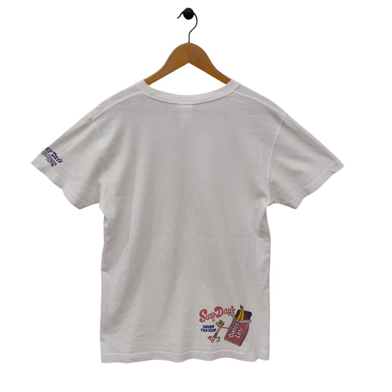 May include: White t-shirt with short sleeves, hanging on a wooden hanger. The shirt features a graphic on the lower right that says "Surf Days" with a cartoon character and a chocolate bar. The left sleeve has text that says "er Days".