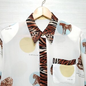 May include: White long-sleeved button-up shirt with a brown and black animal print collar and a pocket. The shirt has a repeating pattern of circles with animal silhouettes and text that reads "Forget me" and "To take me as thy dry dust".