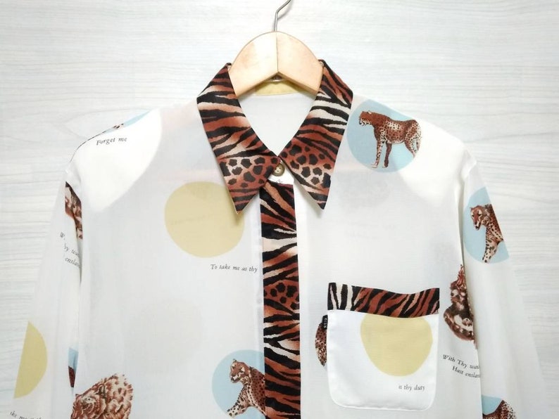May include: White long-sleeved button-up shirt with a brown and black animal print collar and a pocket. The shirt has a repeating pattern of circles with animal silhouettes and text that reads "Forget me" and "To take me as thy dry dust".