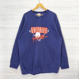 Könnte beinhalten: Ein marineblauer Sweatshirt mit einem weißen Softball und dem Text "Softball Fullerton" in Rot und Weiß.