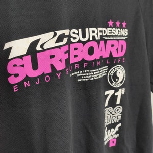 May include: Black t-shirt with white and pink text that reads "T&C Surf Designs", "Surfboard", "Enjoy Surfin' Life", "71#", "T&C Surf", and "Surf".