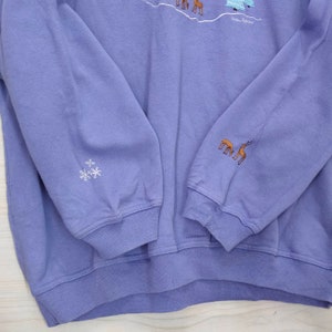 May include: Purple sweatshirt with a winter scene embroidered on the front. The scene includes trees, snow, and deer. The sweatshirt has a snowflake embroidered on one sleeve and a group of deer on the other sleeve.