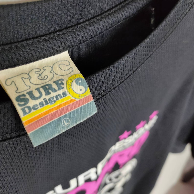 May include: Black t-shirt with a pink and white graphic design that says "Surf Designs" and a white tag with the text "T&C Surf Designs" and a yin yang symbol.