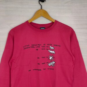 May include: A red sweatshirt with a white graphic print of Keith Haring's drawings. The text reads "Little drawings of Keith Haring characters. Like this or this or this or this or whatever you want."