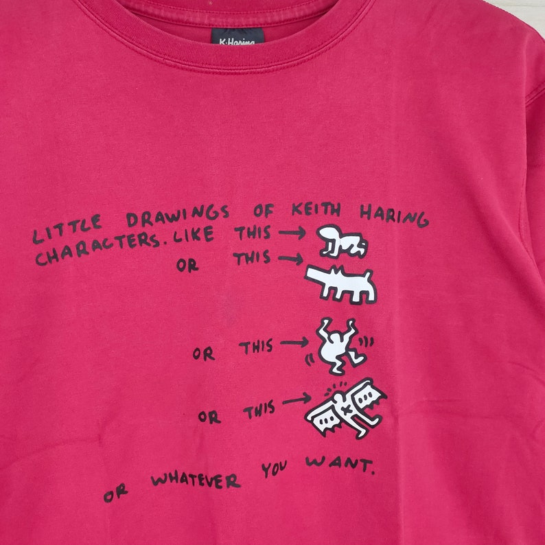 May include: A red t-shirt with a black graphic print of Keith Haring's drawings. The text reads "LITTLE DRAWINGS OF KEITH HARING CHARACTERS. LIKE THIS OR THIS OR THIS OR THIS OR WHATEVER YOU WANT."