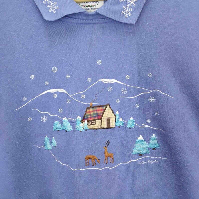 May include: A purple sweatshirt with an embroidered design of a cabin in a snowy mountain landscape. The design includes two deer and the text "Northern Reflection".