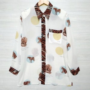 May include: A white button-down shirt with a long sleeve and a leopard print pattern. The shirt has a pocket on the left side and a collar with a zebra print trim. The shirt is hanging on a wooden hanger.