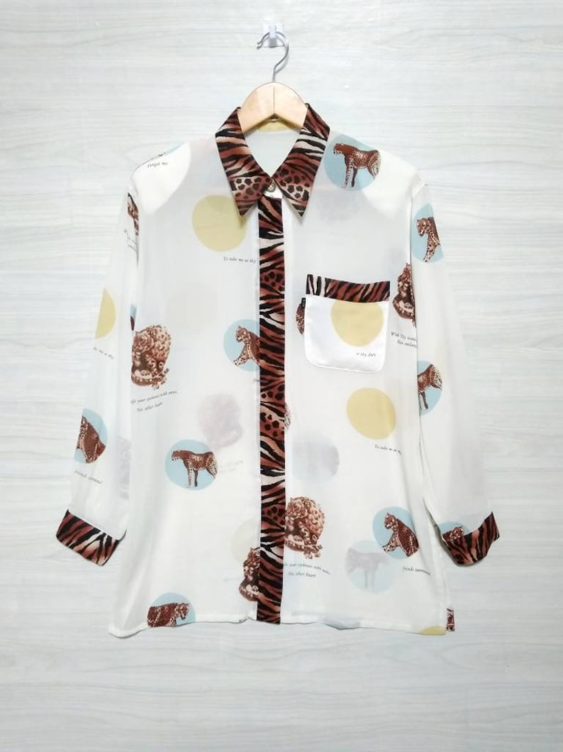 May include: A white button-down shirt with a long sleeve and a leopard print pattern. The shirt has a pocket on the left side and a collar with a zebra print trim. The shirt is hanging on a wooden hanger.