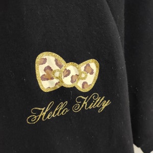 May include: Black garment featuring a gold glitter bow with a leopard print design. The text "Hello Kitty" is embroidered in gold script below the bow. The bow is centered on the garment.