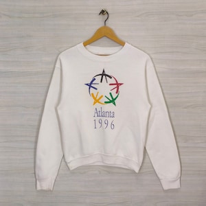 Peut inclure: Sweat-shirt blanc avec un graphique coloré du logo des Jeux olympiques d'Atlanta 1996. Le logo représente une étoile stylisée à cinq branches, chacune représentant une couleur différente des anneaux olympiques. Le texte "Atlanta 1996" est imprimé sous le logo.