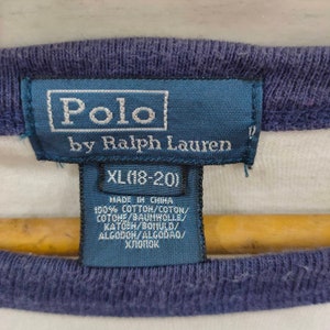 May include: A white and blue striped Polo by Ralph Lauren t-shirt tag with a blue border. The tag says "Polo by Ralph Lauren" and "XL (18-20)". The tag also lists the materials and origin of the shirt: "Made in China, 100% Cotton/Coton/Cotone/Baumwolle/Katoen/Bomuld/Algodon/Algodao/Хлопок."