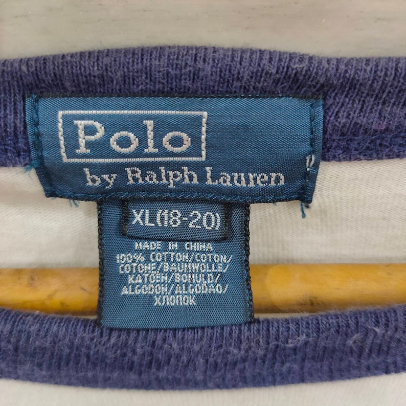 May include: A white and blue striped Polo by Ralph Lauren t-shirt tag with a blue border. The tag says "Polo by Ralph Lauren" and "XL (18-20)". The tag also lists the materials and origin of the shirt: "Made in China, 100% Cotton/Coton/Cotone/Baumwolle/Katoen/Bomuld/Algodon/Algodao/Хлопок."