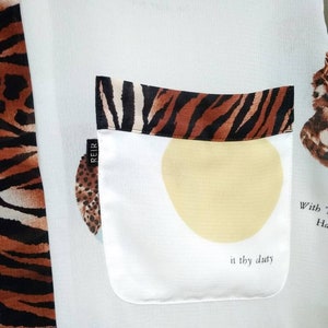 May include: White shirt with a tiger print on the left side and a pocket on the right side. The pocket has a tiger print trim and a yellow circle with the text "in thy duty" inside. The shirt also has a small black label with the text "REIR" on it.