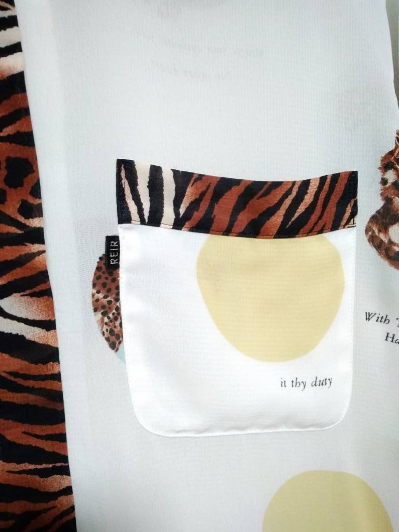 May include: White shirt with a tiger print on the left side and a pocket on the right side. The pocket has a tiger print trim and a yellow circle with the text "in thy duty" inside. The shirt also has a small black label with the text "REIR" on it.