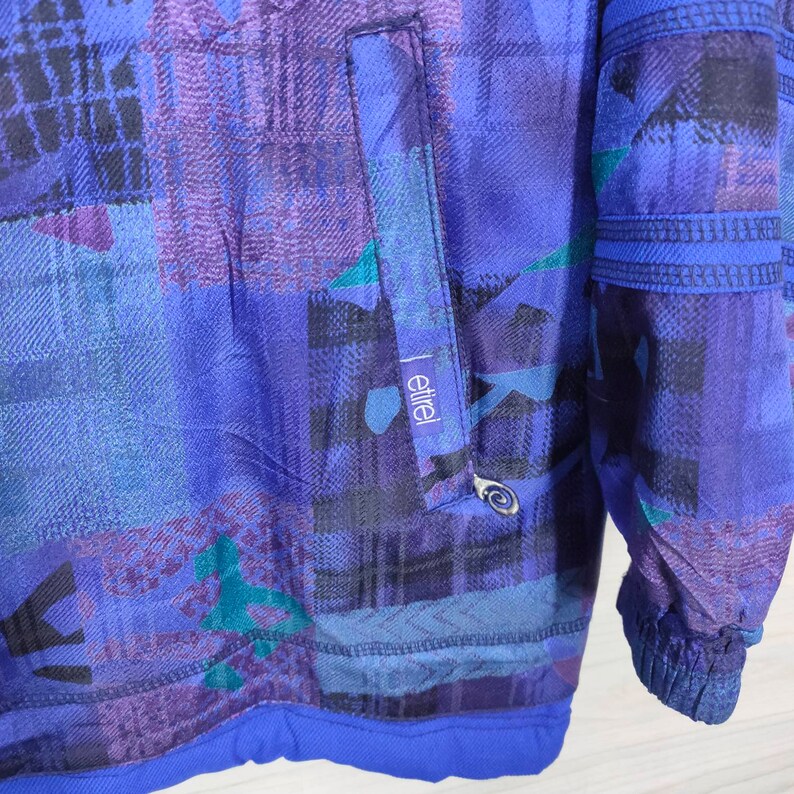 Puede incluir: Una chaqueta de color azul y morado con estampado, cierre de cremallera y un bolsillo en el lado izquierdo. La chaqueta tiene la palabra "etriel" impresa en el bolsillo.
