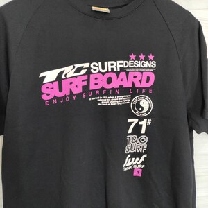 May include: Black t-shirt with white and pink text that reads "TG SURFDESIGNS SURFBOARD ENJOY SURFIN' LIFE" and a yin-yang symbol with the text "T&C Surf Designs Hawaii 71# T&C SURF surf T&C SURF".