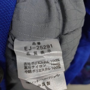 Puede incluir: Una etiqueta de tela gris con texto negro que dice "品番 EJ-25291 品質表示 表地ポリエステル 100% 裏地ナイロン 100% 中綿 ポリエステル 100% (株)インタースポーツジャパン 中国製".