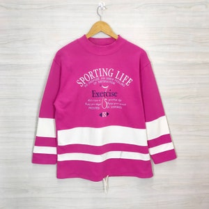 Puede incluir: Una sudadera rosa con rayas blancas en las mangas. La sudadera tiene un gran estampado gráfico en la parte delantera que dice "Sporting Life" y "Exercise".