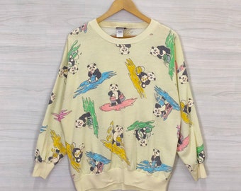 May include: A yellow long-sleeved sweatshirt with a panda bear print. The pandas are wearing colorful swimsuits and are riding on surfboards. The background is a light yellow with blue, green, and yellow accents.