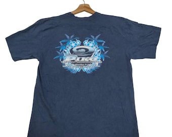 Può includere: T-shirt blu scuro con grafica sul retro. La grafica presenta la parola "PIKO" in un font stilizzato, circondata da disegni floreali e di foglie blu. La maglietta è appesa a una gruccia di legno.