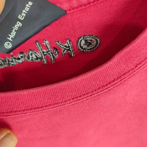 May include: Close-up of a red t-shirt with a black embroidered logo that says "Keith Haring" and a small black and white circle logo that says "&copy; Haring Estate".