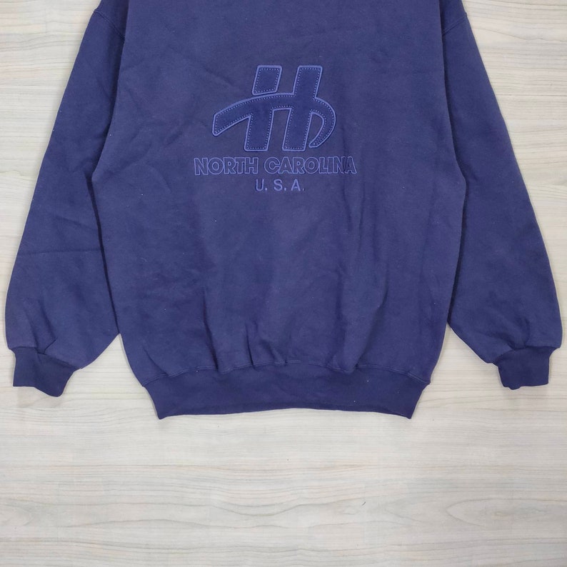 Pode incluir: Uma camisola azul marinho com um grande logotipo bordado que diz "# NORTH CAROLINA U.S.A."