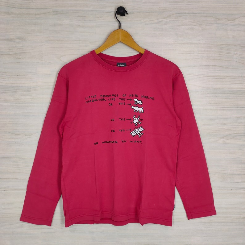 May include: A red long-sleeved t-shirt with a white graphic print of Keith Haring's drawings. The text on the shirt reads "LITTLE DRAWINGS OF KEITH HARING CHARACTERS. LIKE THIS OR THIS OR THIS OR THIS OR WHATEVER YOU WANT."