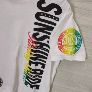 Puede incluir: Camiseta blanca con la palabra "SUNSHINE" en letras negras grandes en el lateral, con dise&ntilde;o de estrellas y rayas. La manga presenta un gr&aacute;fico circular con el texto "BEST SURF WAVE" en degradado arco&iacute;ris.