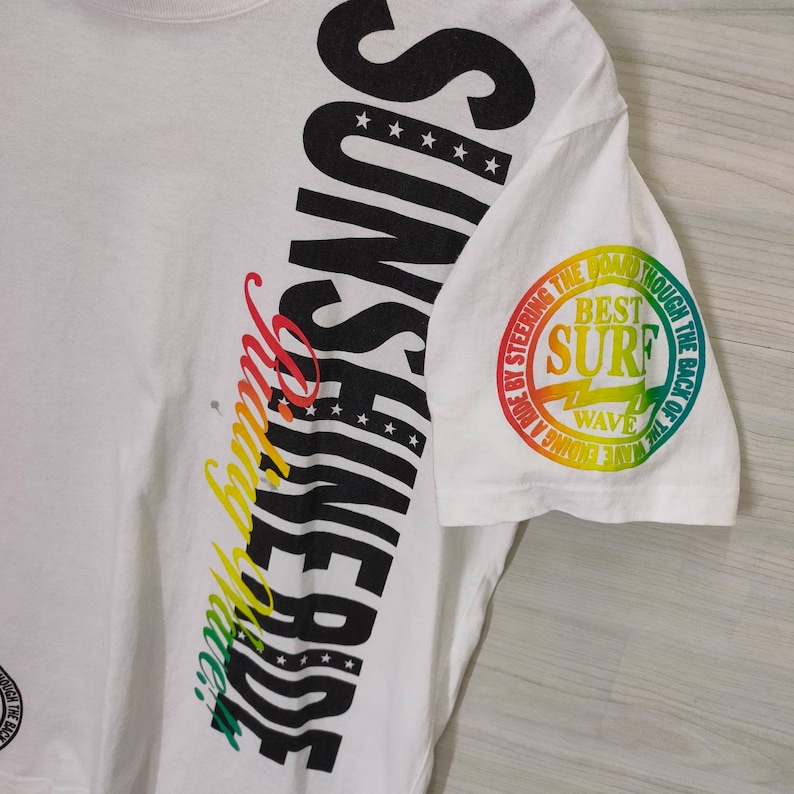 Puede incluir: Camiseta blanca con la palabra "SUNSHINE" en letras negras grandes en el lateral, con dise&ntilde;o de estrellas y rayas. La manga presenta un gr&aacute;fico circular con el texto "BEST SURF WAVE" en degradado arco&iacute;ris.