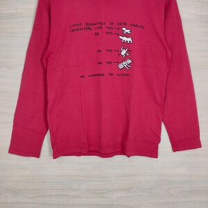 May include: A red long-sleeved shirt with a white graphic print of Keith Haring characters. The text on the shirt reads "LITTLE DRAWINGS OF KEITH HARING CHARACTERS. LIKE THIS OR THIS OR THIS OR THIS OR WHATEVER YOU WANT."