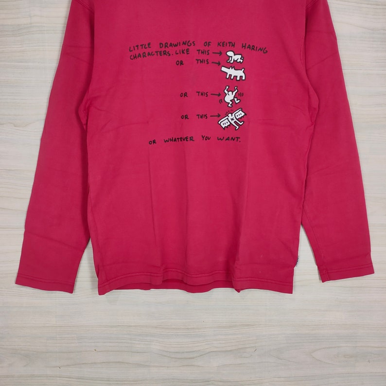 May include: A red long-sleeved shirt with a white graphic print of Keith Haring characters. The text on the shirt reads "LITTLE DRAWINGS OF KEITH HARING CHARACTERS. LIKE THIS OR THIS OR THIS OR THIS OR WHATEVER YOU WANT."