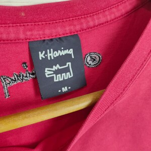 May include: A red t-shirt with a black K. Haring label featuring a white graphic of a dog and the text "K. Haring" and "- M -" .