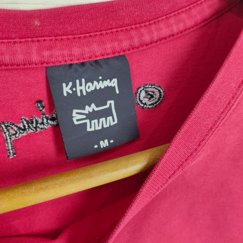 May include: A red t-shirt with a black K. Haring label featuring a white graphic of a dog and the text "K. Haring" and "- M -" .