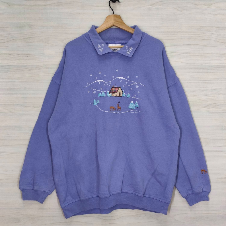 May include: A purple sweatshirt with a winter scene embroidered on the front. The scene includes a small house, trees, and two deer. The collar is trimmed with white snowflakes.