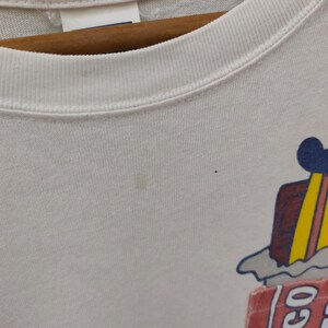 May include: White crew neck t-shirt with a colorful graphic on the lower right. The graphic includes a red sign with the word "GO" and a cartoon image of a building with a blue roof and yellow stripes.