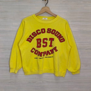 Puede incluir: Sudadera amarilla con un estampado gráfico rojo y negro que dice "Disco Sound BST Company" con el texto "Paris - Tokyo - Los Angeles" debajo.