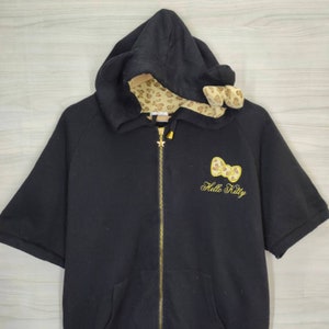 May include: Black zip-up hooded sweatshirt with short sleeves. The hood lining and bow detail are patterned with a tan leopard print. A gold-colored Hello Kitty bow and text are embroidered on the front. The zipper has a star-shaped pull.