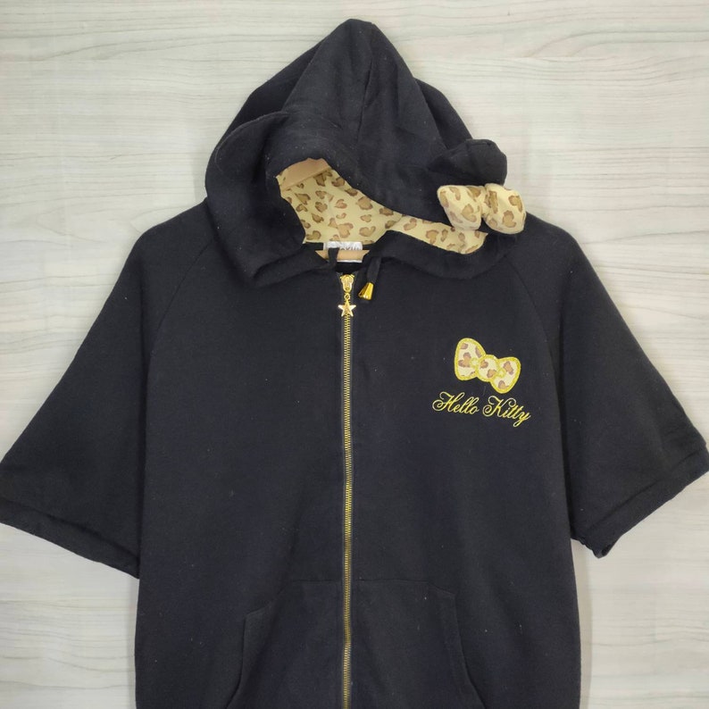 May include: Black zip-up hooded sweatshirt with short sleeves. The hood lining and bow detail are patterned with a tan leopard print. A gold-colored Hello Kitty bow and text are embroidered on the front. The zipper has a star-shaped pull.