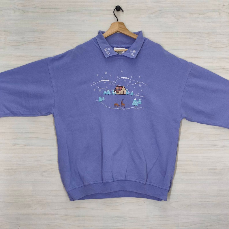 May include: A purple sweatshirt with a winter scene embroidered on the front. The scene includes a small house, trees, and deer. The collar is trimmed with white snowflakes.