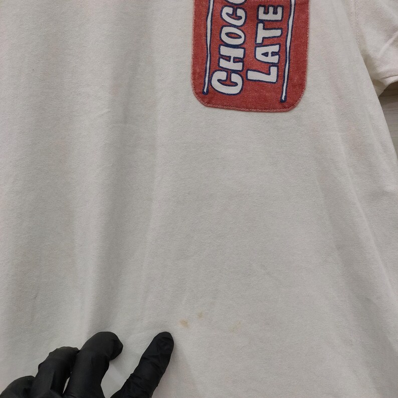May include: Cream-colored t-shirt with a red pocket featuring the words "CHOCOLATE" and "LATE" in white and blue. The shirt has short sleeves and a slightly worn appearance.