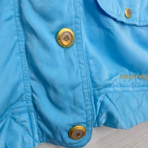 May include: Close-up of a light blue jacket with two gold buttons featuring a tree logo. The text "Courreges" is embroidered on the jacket.