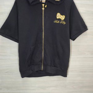 May include: Black short-sleeved zip-up hoodie with a gold zipper and a hood. The hoodie features a gold embroidered Hello Kitty bow and text on the front. The hoodie has two front pockets and a star charm on the zipper.