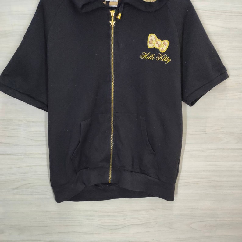 May include: Black short-sleeved zip-up hoodie with a gold zipper and a hood. The hoodie features a gold embroidered Hello Kitty bow and text on the front. The hoodie has two front pockets and a star charm on the zipper.