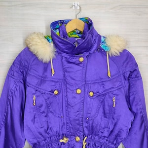 May include: A purple ski jacket with a white fur-lined hood and gold buttons. The jacket has a drawstring waist and zippered pockets.