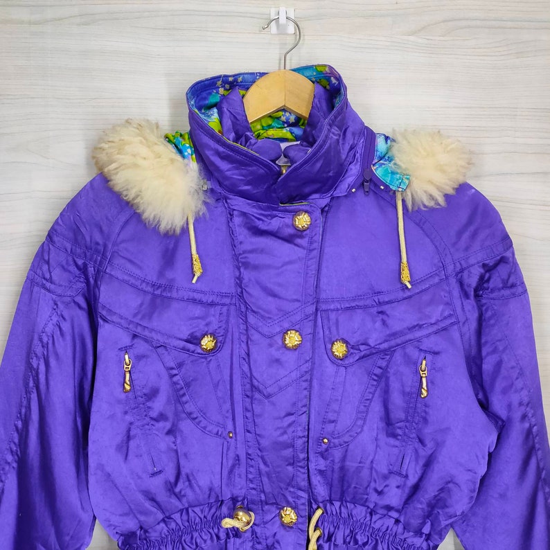 May include: A purple ski jacket with a white fur-lined hood and gold buttons. The jacket has a drawstring waist and zippered pockets.
