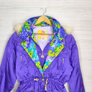 May include: A purple ski jacket with a floral lining and a fur-trimmed hood. The jacket has a zipper closure, a drawstring waist, and gold-toned hardware. The label on the jacket reads "Ellen Tracy".